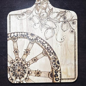 Peut inclure: Planche à découper en bois avec une poignée, ornée de motifs pyrogravés complexes. La planche présente un motif floral et de citron, ainsi qu'un motif de roue décorative. Les bords sont ornés d'un motif en forme de chaîne, créant une esthétique rustique.