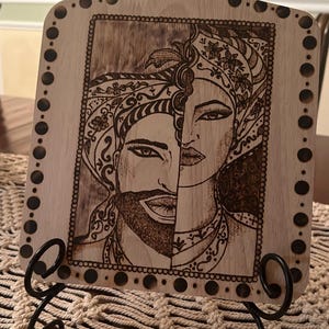 May include: A wooden cutting board with a decorative design of two faces, one male and one female, burned into the surface. The board has a handle with a hole in the top and a decorative border of black dots.