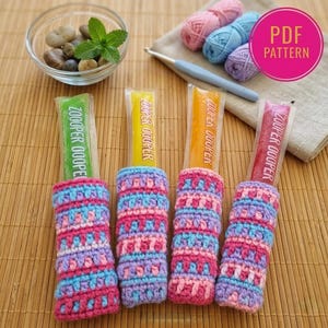 Puede incluir: Cuatro helados Zooper Dooper en fundas de ganchillo hechas a mano. Las fundas son rosas, azules y moradas. La imagen también muestra lana, un ganchillo y un pequeño cuenco con piedras y menta.