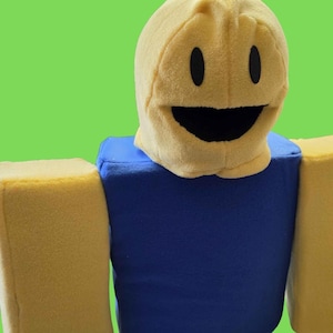 May include: A plush toy character featuring a yellow rounded head with black oval eyes and a wide black mouth. The torso is a solid blue rectangular block, and the two arms are solid yellow rectangular blocks, all made of soft fabric.