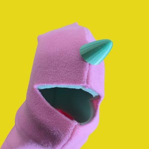 May include: A pink fabric puppet head with a green horn and a large open mouth. The puppet has a simple design and is set against a bright yellow background. The mouth interior is dark.