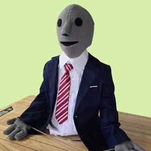 May include: A gray puppet wearing a blue suit with a red and white striped tie. The puppet is sitting on a wooden surface with a ruler and protractor.
