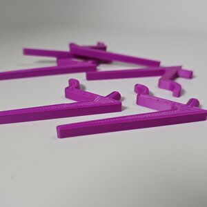 May include: A collection of bright purple, 3D-printed shelf brackets. The brackets have a right-angle design with a curved hook on one end. They are scattered on a white surface, with some in focus and others blurred.