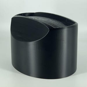 Puede incluir: Un recipiente de color negro, de forma ovalada, impreso en 3D, con una parte superior lisa y redondeada y un borde ligeramente hundido.
