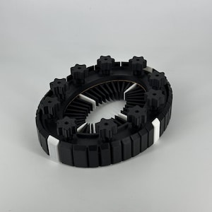 Op de afbeelding: Een zwart-wit cirkelvormig object met stervormige knoppen. Het object heeft een reeks zwarte rechthoekige blokken rond de omtrek, met witte accenten. Het midden heeft een reeks witte vinnen.
