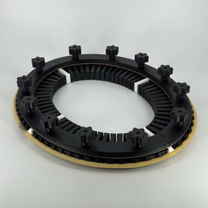 May include: A circular black object with black knobs around the top. It has a beige outer ring and white sections. The object is set against a white background.
