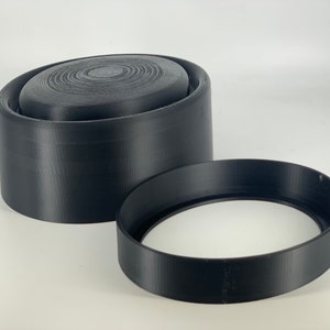 Op de afbeelding: Twee zwarte plastic ringen, een groter dan de andere, met een glad, licht getextureerd oppervlak. De grotere ring heeft een plat, rond bovenoppervlak.