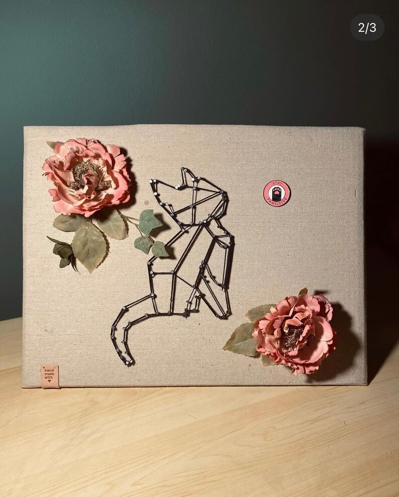May include: A black wire cat sculpture on a beige canvas background with two pink fabric flowers. The canvas has a small brown label that says "hand made with love". There is a pink circle sticker with a black paw print and the text "Stay Awesome".