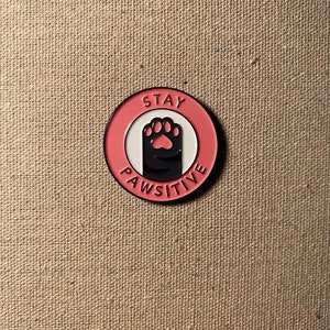 May include: Pink and white enamel pin with a black paw print and the text "Stay Pawsitive".