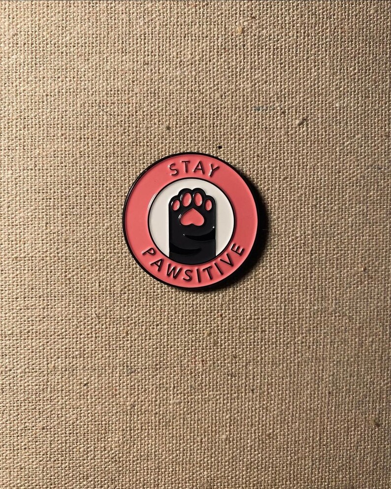 May include: Pink and white enamel pin with a black paw print and the text "Stay Pawsitive".