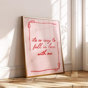May include: A framed print with the text "its so easy to fall in love with me" in red script. The print has a pink background and a gold frame. The frame is leaning against a white wall and wooden floor.
