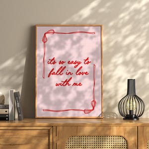 May include: A framed print with the text "it's so easy to fall in love with me" in red script on a pink background. The frame is a light brown color. A decorative black lamp with a light bulb is on a wooden cabinet.