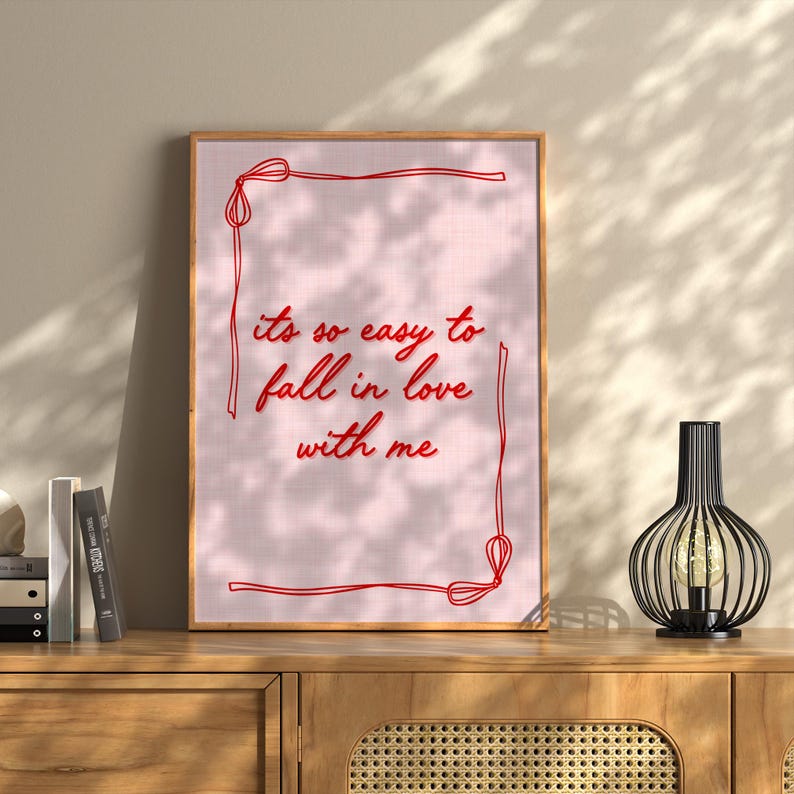 May include: A framed print with the text "it's so easy to fall in love with me" in red script on a pink background. The frame is a light brown color. A decorative black lamp with a light bulb is on a wooden cabinet.
