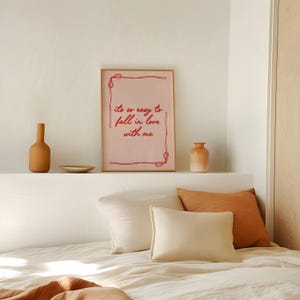May include: A framed pink print with the text "it's so easy to fall in love with me" in red script. The print is displayed above a bed with pillows and decorative vases in shades of orange and beige.