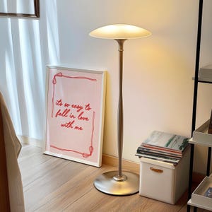 May include: A modern floor lamp with a brushed metal finish and a wide, disc-shaped lampshade. The lamp stands tall next to a framed print with the text "it's so easy to fall in love with me". A white storage box and a shelf are also visible.