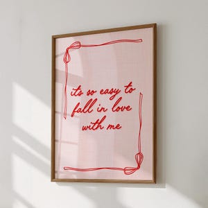 May include: A framed print with a light pink background and the text "its so easy to fall in love with me" in red cursive. The print is framed in a light brown wooden frame, with a red border.