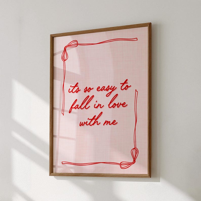 May include: A framed print with a light pink background and the text "its so easy to fall in love with me" in red cursive. The print is framed in a light brown wooden frame, with a red border.