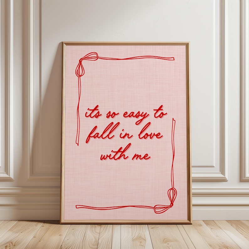 May include: Framed art print with a light pink background and red text that reads "its so easy to fall in love with me." The artwork is framed in a light brown wooden frame and features a red border.