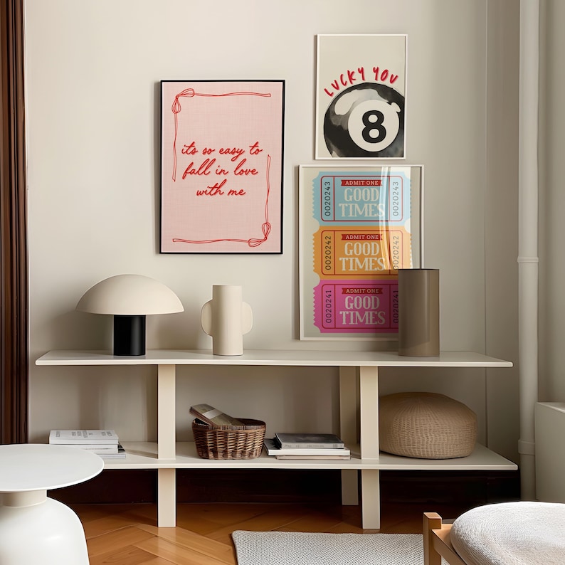 May include: A modern interior scene featuring framed art prints and decorative objects. The prints include text that reads "it's so easy to fall in love with me", a lucky 8-ball, and "GOOD TIMES" tickets. A mushroom-shaped lamp, vase, and woven basket are on a white shelf.