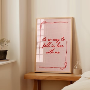 May include: A framed print with the text "it's so easy to fall in love with me" in red script. The print has a pink background and a red border. The frame is made of light-colored wood. The print is displayed on a wooden table.