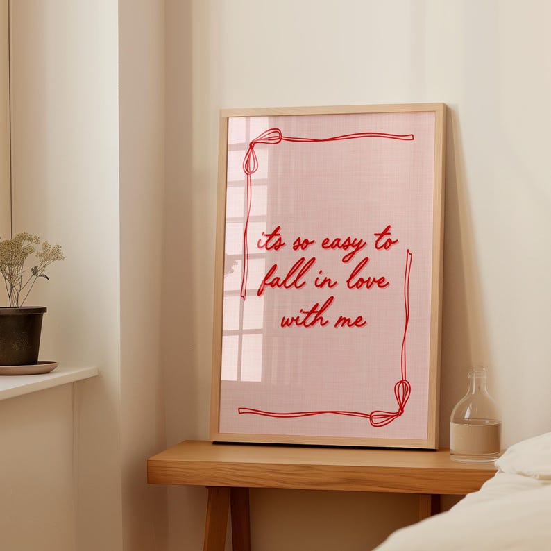 May include: A framed print with the text "it's so easy to fall in love with me" in red script. The print has a pink background and a red border. The frame is made of light-colored wood. The print is displayed on a wooden table.