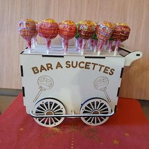 Peut inclure: Un chariot en bois blanc avec le texte "BAR A SUCETTES" contient plusieurs sucettes emballées. Le chariot a des roues décoratives et des illustrations de sucettes. Les sucettes ont des emballages rouges et jaunes.