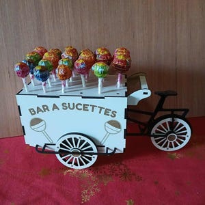 May include: A white wooden cart with a black bicycle frame, holding colorful lollipops. The cart has the text "BAR A SUCETTES" and lollipop illustrations. The lollipops have various wrappers and colors, arranged on white sticks.