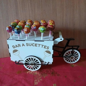 Könnte beinhalten: Ein weißer Holzkarren mit schwarzem Fahrradrahmen, der bunte Lutscher enthält. Der Wagen trägt den Text "BAR A SUCETTES" und Lutscher-Illustrationen. Die Lutscher haben verschiedene Verpackungen und Farben, angeordnet auf weißen Stäben.