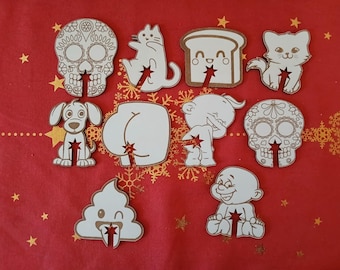 Puede incluir: Colección de figuras de madera cortadas con láser, con acabado blanco y bordes marrones. El conjunto incluye una calavera de azúcar, un gato, un perro, pan y otras formas humorísticas, cada una con un recorte en forma de estrella. Sobre fondo rojo.