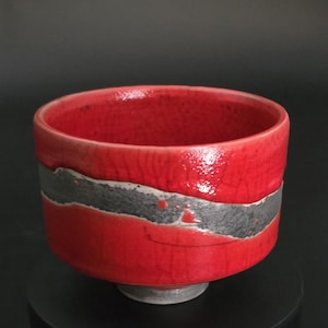 Könnte beinhalten: Eine rote Keramikschale mit einem breiten, dunkelgrauen Band in der Mitte. Die Schale hat eine glänzende Oberfläche und steht auf einem kleinen, runden Sockel. Auch das Innere der Schale ist rot.