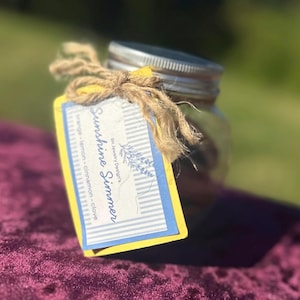 Könnte beinhalten: Ein klares Glasgefäß mit einem silbernen Metalldeckel, an dem ein rechteckiges Etikett mit einer Schnur befestigt ist. Das Etikett hat einen gelben Rand, blau-weiße Streifen und den Text Sunshine Simmer, BK Jewelry Design's sowie orange - lemon - cinnamon - clove.