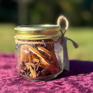 May include: A small clear glass jar with a gold metal lid, filled with dried orange peels, star anise, and cinnamon sticks. A piece of natural jute twine is wrapped around the neck of the jar, and a small beige tag hangs from the side.