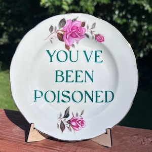 May include: A white ceramic plate with a scalloped edge and a metallic silver rim. The center features the dark green text "YOU'VE BEEN POISONED" with pink rose illustrations and green leaves positioned above and below the printed message.
