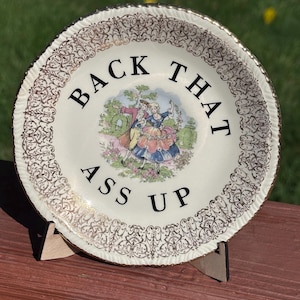 May include: A decorative ceramic plate with a scalloped edge and gold filigree border. The center features a colorful illustration of two figures in historical attire dancing, surrounded by the black text "BACK THAT ASS UP" in a circular layout.