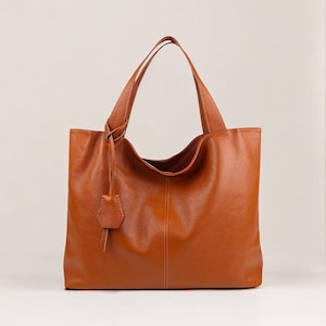 Peut inclure: Un grand sac fourre-tout rectangulaire en cuir marron avec deux anses. Le sac a une surface texturée et une petite étiquette décorative en cuir attachée à la poignée. Le sac est vide et se trouve sur un fond uni.