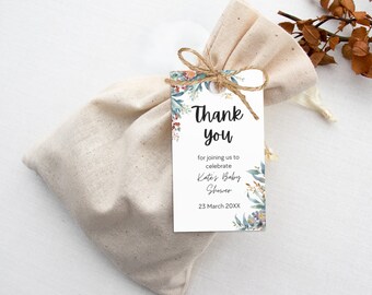 Puede incluir: Una etiqueta blanca de agradecimiento con un diseño floral y el texto "Gracias por unirse a nosotros para celebrar el Baby Shower de Kate 23 de marzo de 20XX". La etiqueta está atada a una pequeña bolsa de tela blanca.