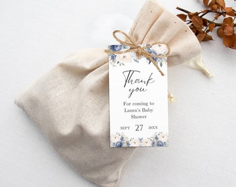 Op de afbeelding: Een beige stoffen cadeauzakje vastgebonden met touw, met een klein item. Een wit label met blauwe en crèmekleurige bloemmotieven en de woorden "Thank you" is bevestigd. Het label bevat ook de tekst "For coming to Laura's Baby Shower".