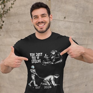 Puede incluir: Camiseta negra con gráficos con el texto "YOU JUST HAD TO EVOLVE" e ilustraciones de un mono, un robot y una persona. La camiseta también tiene impreso "EST. 2026".
