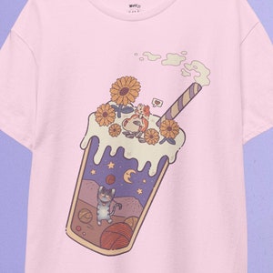 May include: A pink t-shirt featuring a cartoon illustration of a glass of iced coffee with a straw, flowers, and a cat inside. The coffee is topped with whipped cream and a cherry.