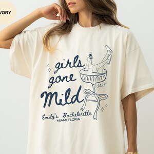 May include: Ivory oversized t-shirt with a graphic of a woman in a champagne glass. The text on the shirt reads "girls gone mild" and "Emily's Bachelorette Miami, Florida".