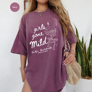May include: Purple oversized t-shirt with the text "girls gone mild" and a graphic of a woman in a champagne glass. The text "Emily's Bachelorette Miami, Florida" is also printed on the shirt.