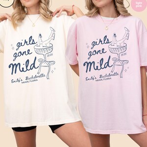 May include: Two oversized t-shirts with a graphic design of a woman in a martini glass with the text "girls gone mild" and "Emily's Bachelorette Miami, Florida 2025". The t-shirts are in ivory and light pink.