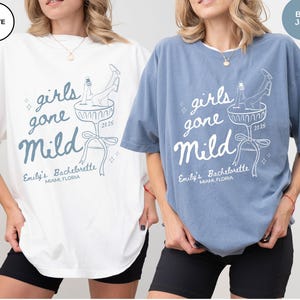 May include: Two oversized t-shirts with a white graphic design that says "girls gone mild" with a cocktail glass and a woman's leg sticking out of the glass. The text "Emily's Bachelorette Miami, Florida 2025" is below the graphic. The t-shirt on the left is white and the t-shirt on the right is blue jean.