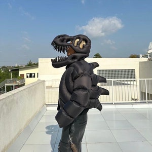 Könnte beinhalten: Ein schwarzes Dinosaurierkostüm, bestehend aus einer gepolsterten Jacke mit klauenartigen Verlängerungen und einer Dinosaurierkopfhaube. Das Kostüm wird im Freien vor dem Hintergrund eines blauen Himmels und weißer Gebäude getragen.