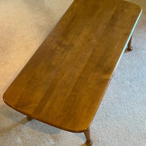 May include: A rectangular wooden coffee table with rounded corners and four curved legs. The table has a warm, medium-brown finish, showcasing the natural wood grain. The table is approximately 48 inches long and 24 inches wide.