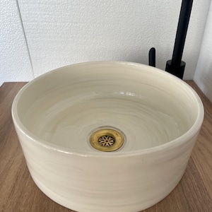 May include: Cream-coloured ceramic vessel sink with a brass drain and a black tap. The sink has a round shape and a smooth, glossy finish. The tap is a modern design.