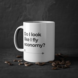 Pode incluir: Caneca de café de cerâmica branca com texto preto que diz "Do I look like I fly economy?" com um pequeno ícone de avião e uma linha pontilhada.