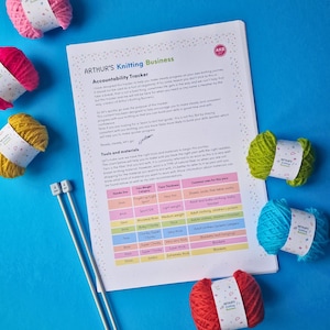 May include: A white sheet of paper with the title "Arthur's Knitting Business" and a table listing yarn weights, yarn thicknesses, and common uses for each yarn. The paper is surrounded by balls of yarn in various colors, including red, yellow, green, and blue.