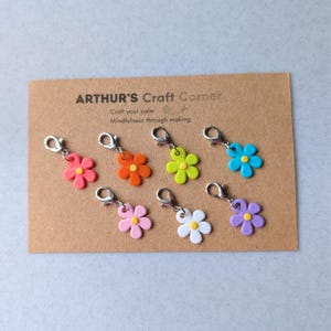 May include: A selection of flower charms in shades of pink, orange, lime green, blue, light pink, white, and purple, each with a silver clasp. They are presented on a brown card with the text "ARTHUR'S Craft Corner".