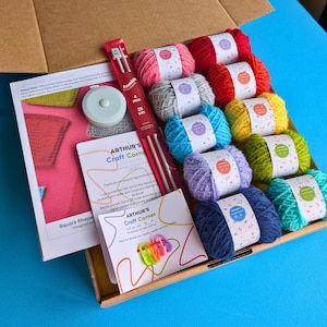 May include: A craft box containing various coloured yarn skeins, including pink, red, grey, blue, yellow, green, purple, and teal. The box also includes knitting needles (4 mm, 25 cm), stitch markers, a tape measure, and printed materials. The box is open on a blue surface.